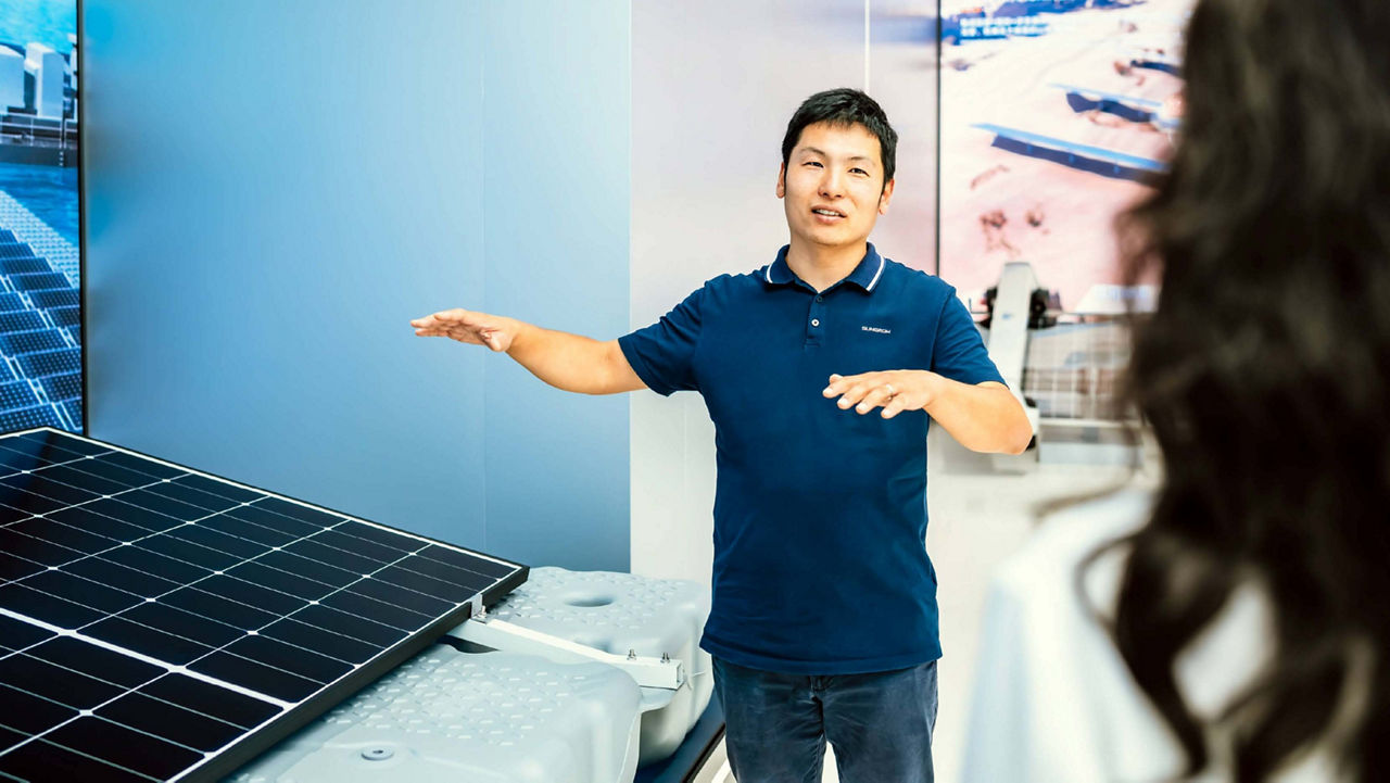 Jie Ding, an Asian man in a Sungrow-branded shirt, is engaged in a product explanation with a woman – she is seen from behind, listening attentively. A professional interaction captured in a modern environment.