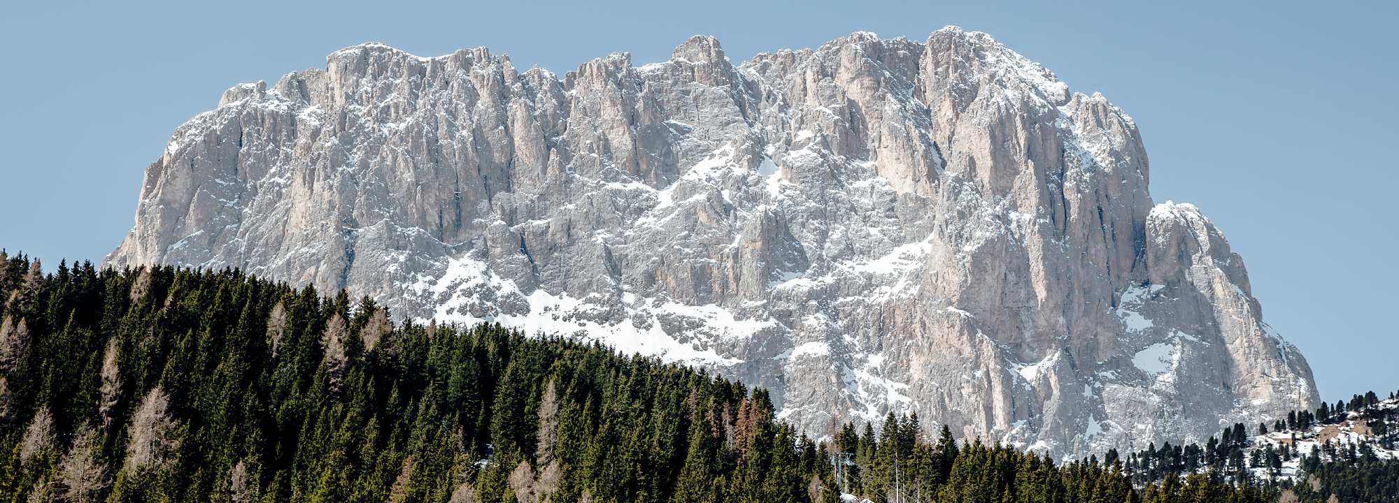 Snow-covered rocky mountain peak rising above a forested hillside. The steep cliff face shows exposed rock formations with patches of snow against a clear blue sky. In the foreground is a dense forest of evergreen trees with some bare trees visible among them.