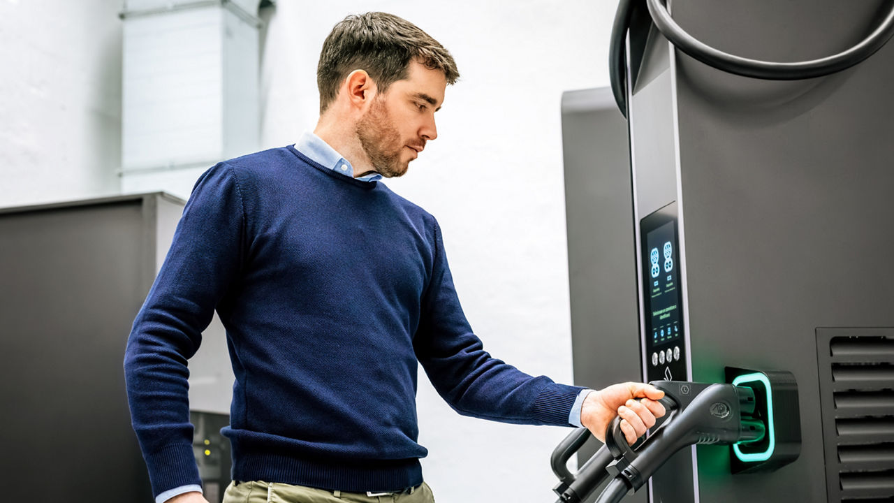 A young man wearing a navy blue sweater over a light blue shirt is standing at an electric vehicle charging station. He is holding a charging plug in his right hand, inserting it into the charging socket of the station. The charging station has a digital display. The background is a white wall.