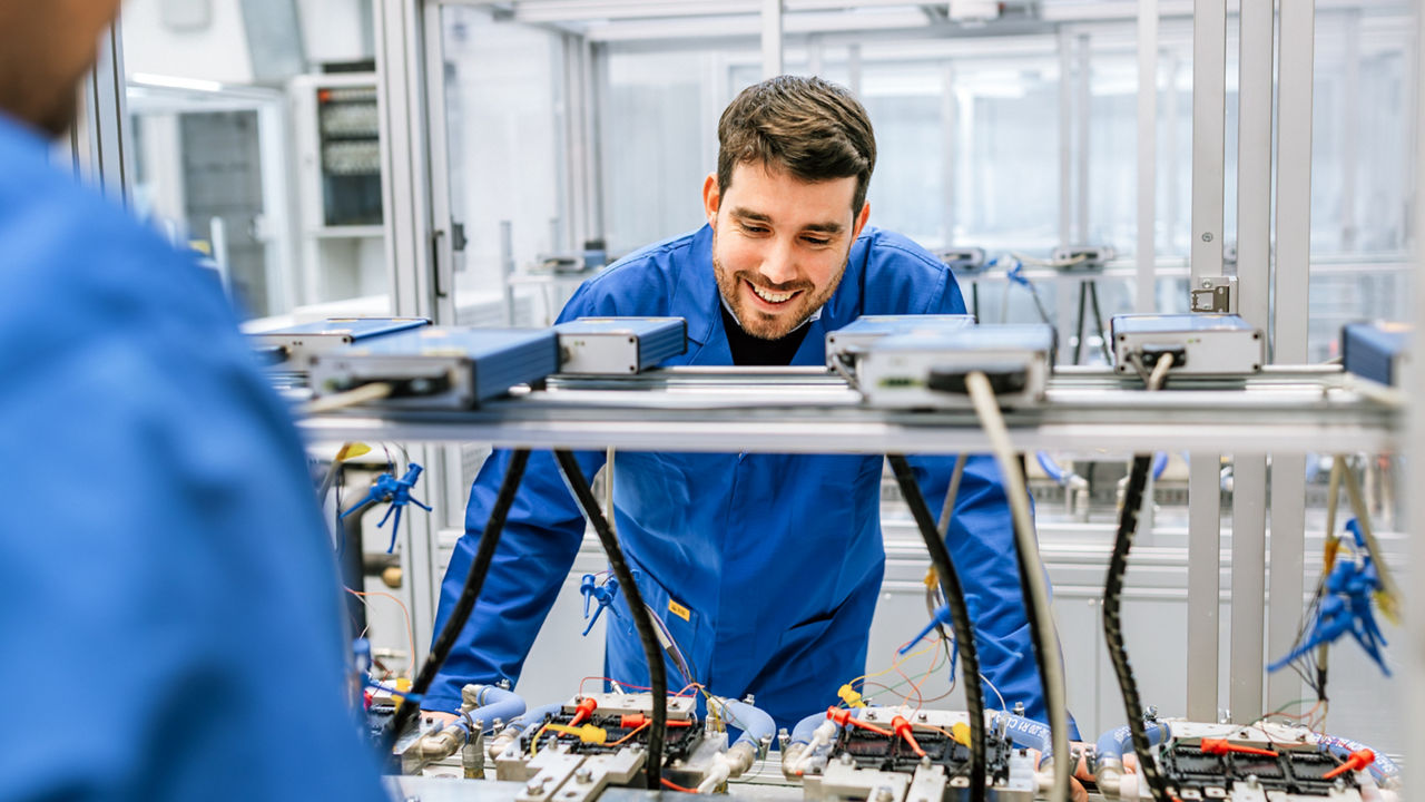 A smiling young man in a blue work coat is examining technical equipment in a laboratory setting. He is leaning over a workstation with multiple blue electronic devices connected by cables on a metal frame. The background shows a clean, modern laboratory environment with glass partitions.