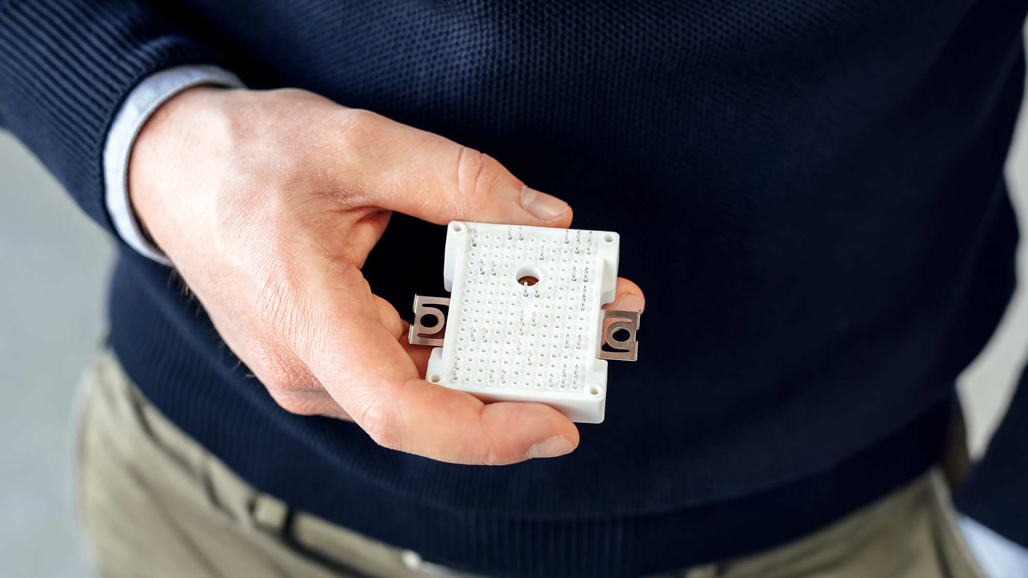 Close-up of a hand holding a small white electronic circuit board or component. The person is wearing a navy blue sweater with a visible light-colored shirt cuff. The circuit board has multiple small points and a central hole, with metal connectors visible on the sides.