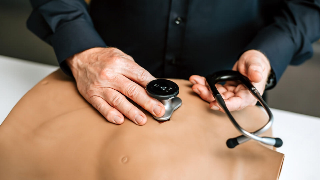 A man in a black shirt demonstrates a smart stethoscope by Eko Health on a test dummy – holding the device to the chest while the headset rests in his other hand