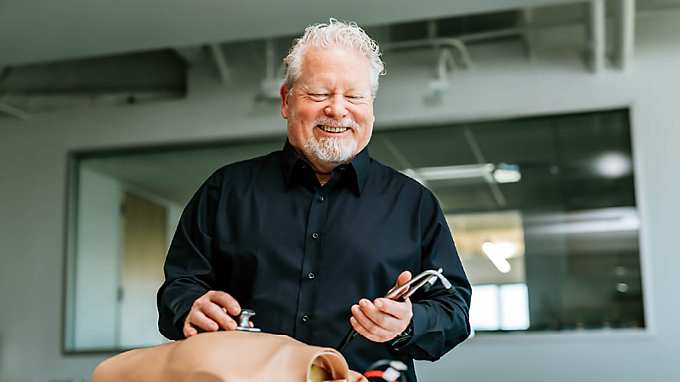 Karl Larsson, wearing a black shirt, stands in a laboratory demonstrating a smart stethoscope by Eko Health on a test dummy. Smiling and focused on the device, he holds the chest piece to the dummy’s chest while the earpiece rests in his other hand – illustrating innovation in digital health diagnostics