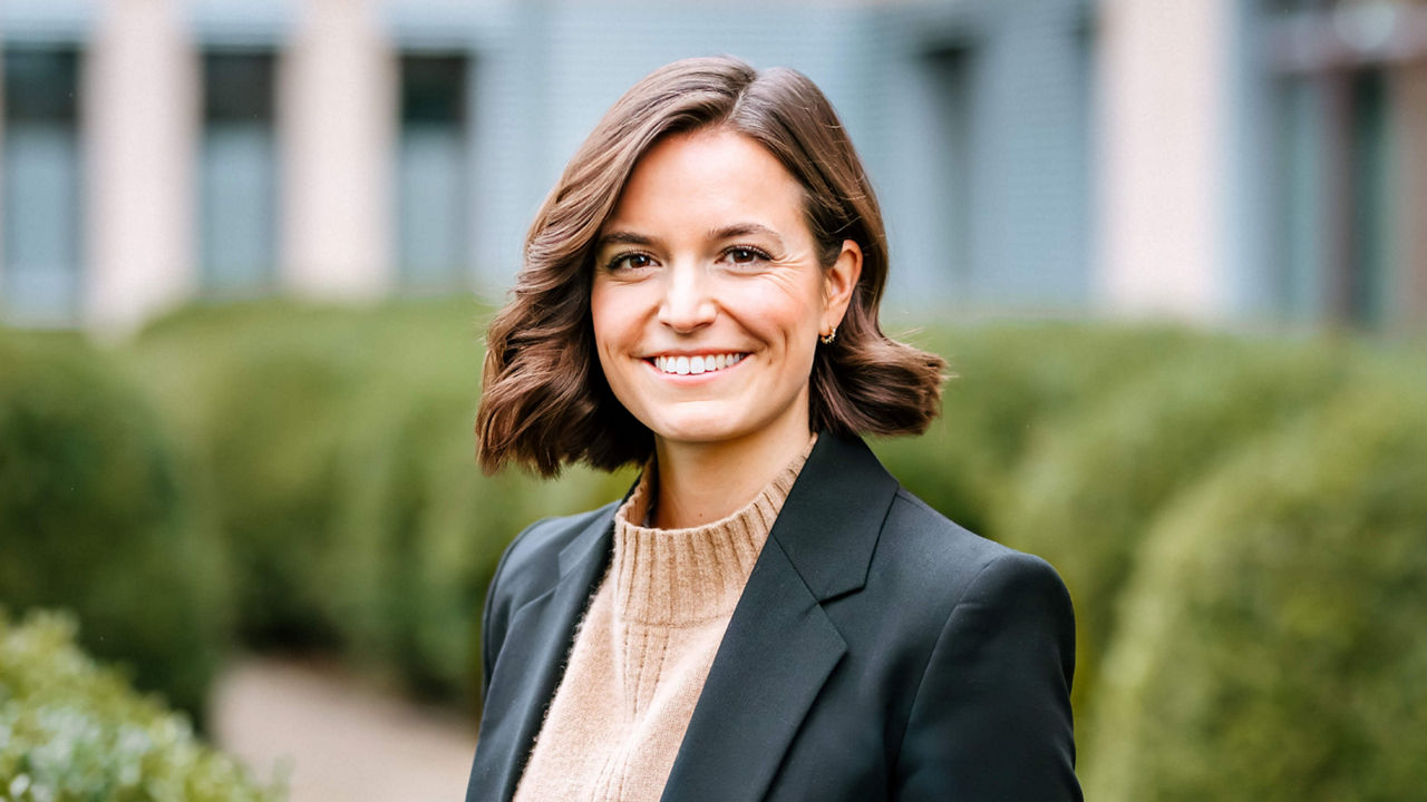 Portrait of Carolin Kerscher. She is smiling in a dark blazer and beige knitted jumper and looks at the camera. Carolin, with wavy brown hair, brown eyes, and a broad smile that shows her teeth, looks very friendly and approachable. She stands in front of the Infineon Campeon building with green bushes in the background.