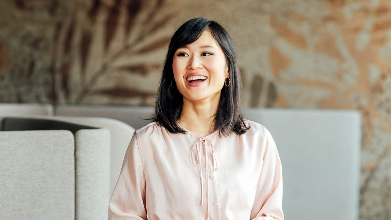 Portrait of Dr. Yinghong Hu in a light pink blouse, laughing and looking to the side. She has shoulder-length black hair with a side fringe, and her eyes are accentuated with dark eyeshadow. She laughs with an open mouth while glancing sideways. Her pink blouse features a button placket and a small bow at the collar. She is wearing gold hoop earrings. The atmosphere is relaxed and cheerful. She is in an office with grey furniture and brown patterned wallpaper.