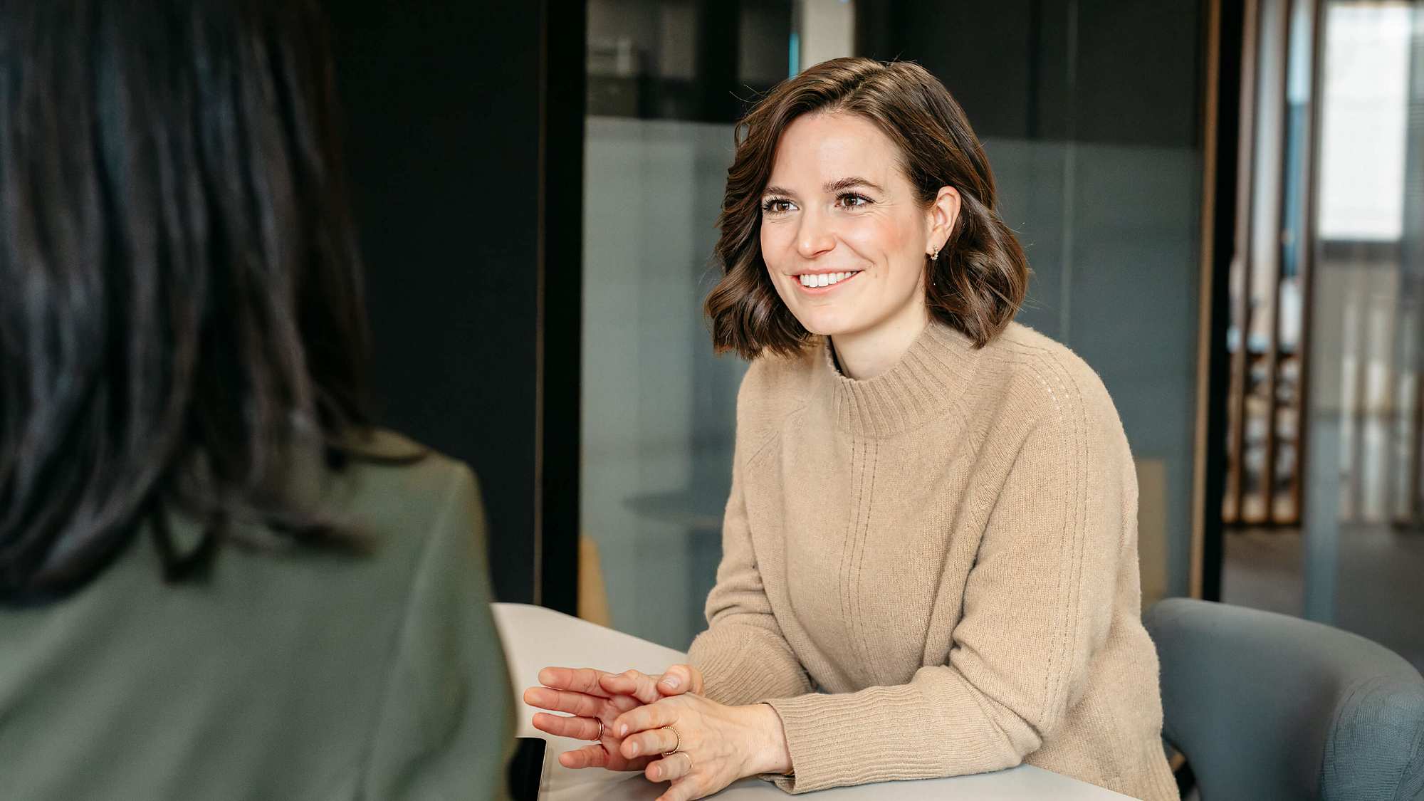 Portrait of Carolin Kerscher interacting with Dr. Yinghong Hu. The focus of the image is on Carolin Kerscher. Only the back of Yinghongs head is visible. Carolin Kerscher is seated at a table, smiling at Yinghong. She is wearing a beige knitted jumper and subtle gold jewelry on her fingers and ears. Her shoulder-length brown wavy hair frames her friendly expression. In the background, a glass phone booth with grey veneer and a wooden panel is visible. The open-plan office reflects a modern and welcoming work atmosphere.