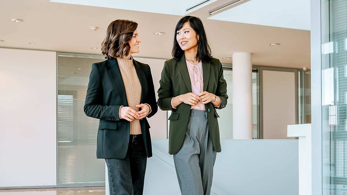 Dr. Yinghong Hu and Carolin Kerscher walk through a large, bright hallway, looking at each other while talking with their hands held in front of their bodies. Carolin is wearing a dark blazer over a beige knitted jumper, paired with dark jeans. She has shoulder-length wavy brown hair. Yinghong is dressed in loose grey suit trousers, a dark green blazer, and a pink blouse with a bow at the collar. She has straight black hair.