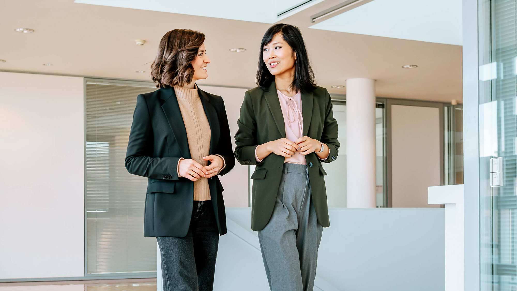 Dr. Yinghong Hu and Carolin Kerscher walk through a large, bright hallway, looking at each other while talking with their hands held in front of their bodies. Carolin is wearing a dark blazer over a beige knitted jumper, paired with dark jeans. She has shoulder-length wavy brown hair. Yinghong is dressed in loose grey suit trousers, a dark green blazer, and a pink blouse with a bow at the collar. She has straight black hair.
