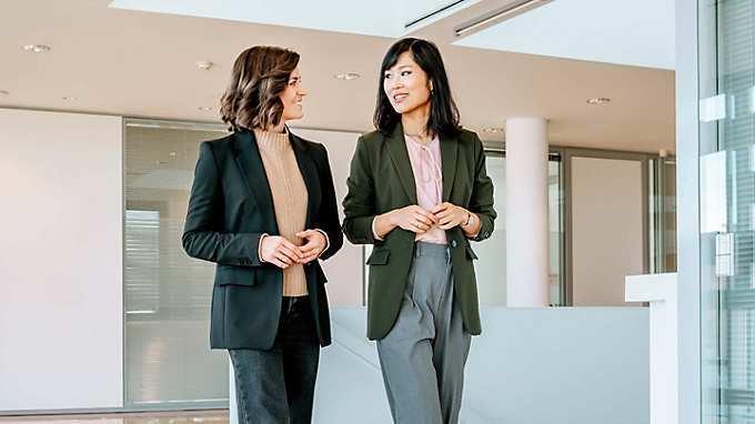 Dr. Yinghong Hu and Carolin Kerscher walk through a large, bright hallway, looking at each other while talking with their hands held in front of their bodies. Carolin is wearing a dark blazer over a beige knitted jumper, paired with dark jeans. She has shoulder-length wavy brown hair. Yinghong is dressed in loose grey suit trousers, a dark green blazer, and a pink blouse with a bow at the collar. She has straight black hair.
