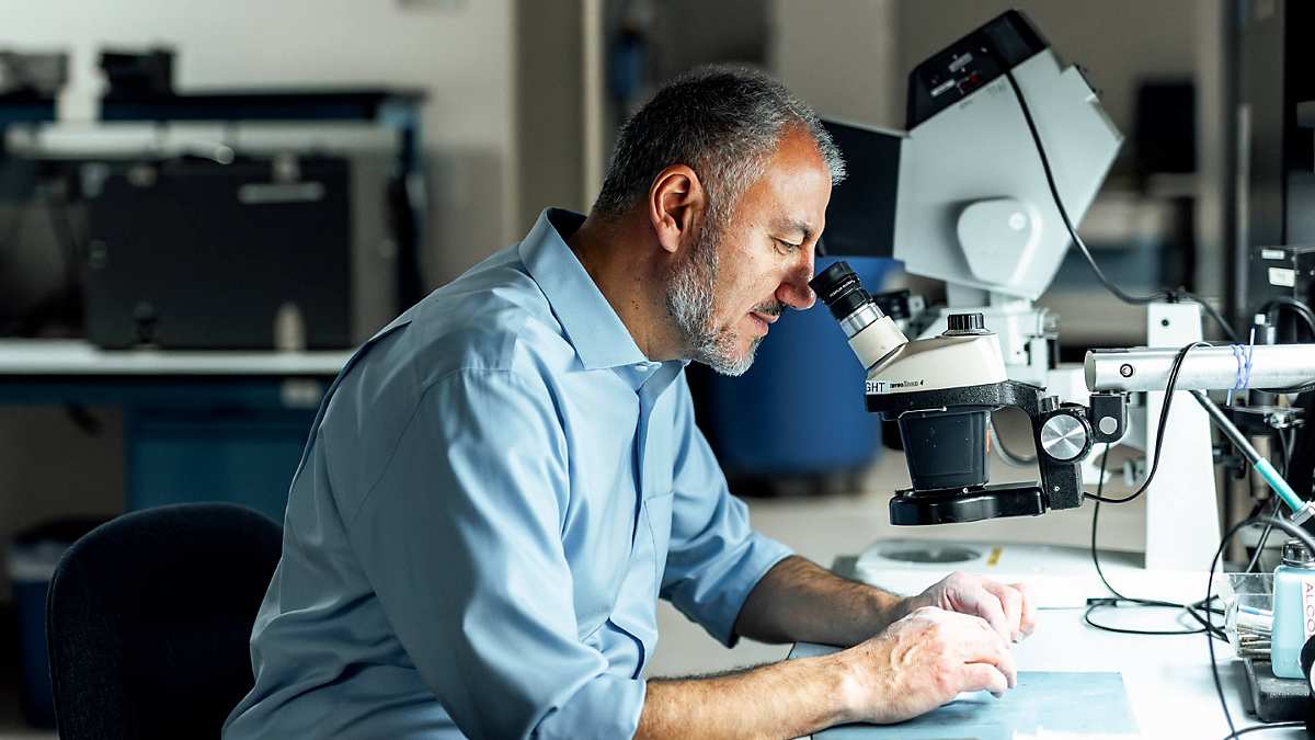 Sam Geha sits at a precision microscope with connected devices, holding his hands under the microscope to closely examine a product. Various tools and items are scattered on the work surface. A light source from the workbench illuminates the desk and Sam's hands. In the background, a blurred view of an engineering lab is visible.