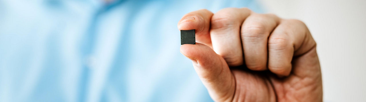 Close-up of a man's hand in a blue shirt, holding a very small Infineon product between thumb and index finger. The remaining fingers are folded in. The back of the product, facing the camera, is black with concentrically arranged solder pads and contacts. The Produkt is called AURIX™ TriCore