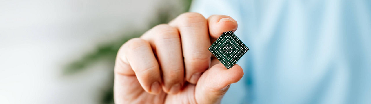 Close-up of a man's hand in a blue shirt, holding a very small Infineon product between thumb and index finger. The remaining fingers are folded in. The black front side of the product is visible, showing a printed product description. The Product is called PSOC™ Edge