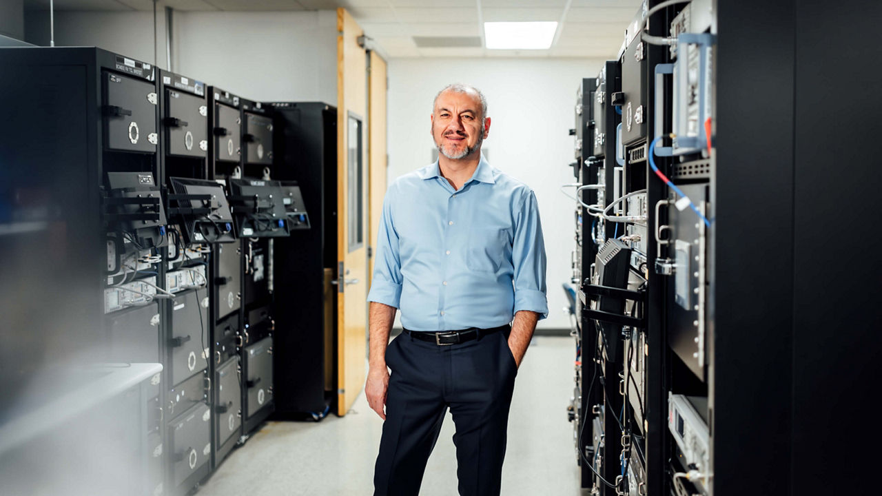 Sam Geha, a bearded man in a light blue shirt and dark trousers, stands casually in the middle of a server room with one hand in his pocket. He is smiling, and black server racks filled with technical equipment line both sides of the room.