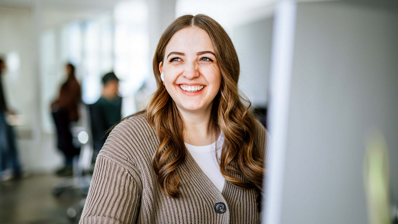 Portrait of Janina smiling and looking to the side. She is wearing white noise-canceling headphones and sitting at a screen in a bright office. In the background, people are working. Janina has long, wavy brown hair and is dressed in a white t-shirt and a brown cardigan.