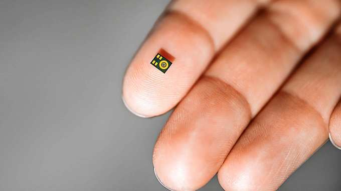 Close-up of a hand in front of a gray background, with a small black microphone resting on the fingertip. The microphone features four yellow rectangles and several concentric yellow circles, highlighting its internal structure or design.