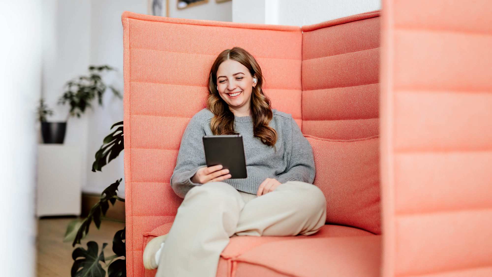 A young woman with long wavy hair sits smiling in a coral-colored high-back sofa. She is wearing a grey sweater and light-colored pants, holding a grey tablet in her hands. She is wearing noise-canceling headphones and appears to be speaking to someone via the tablet. In the background, indoor plants are visible.