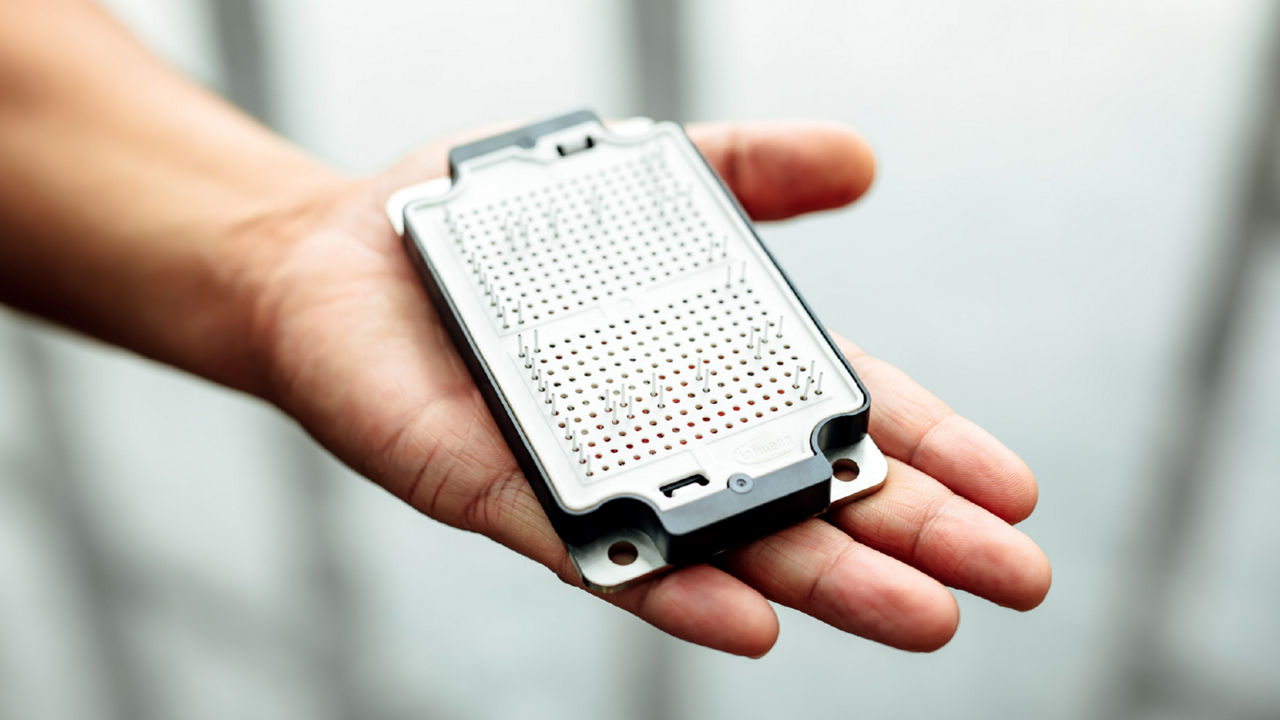 Close-up of a hand holding a white electronic component with a black frame. The circuit board has multiple small points with metal connectors.