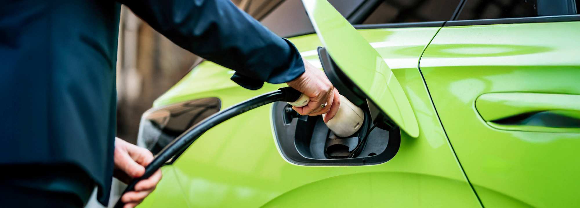 Close-up: Woman in a blue blazer plugs in a green electric car at a charging station. The image captures the moment the connector is being inserted into the car with her right hand, while her left hand holds the cable. The charging port is open. In the background, a mirrored glass office building reflects golden-orange light.