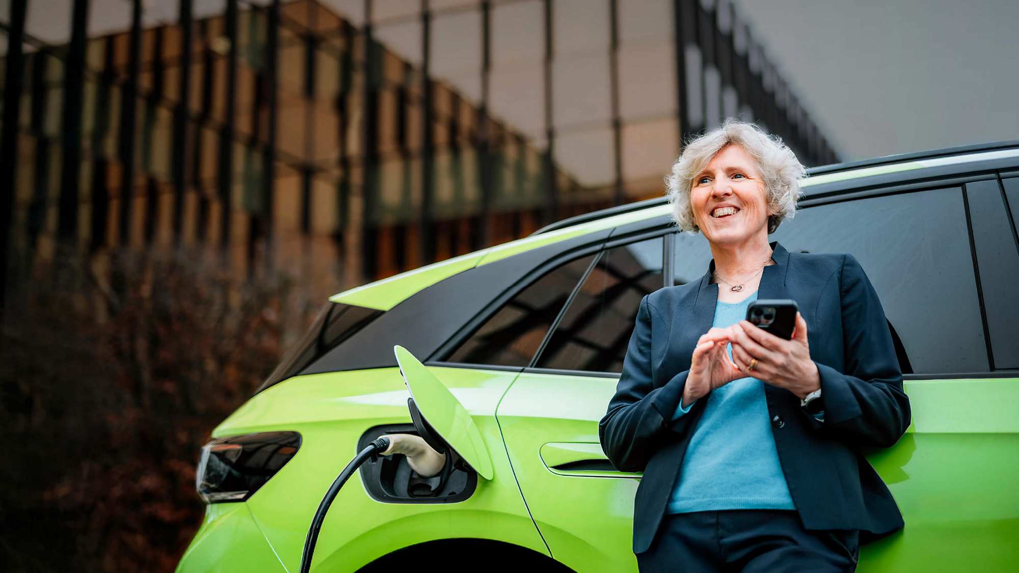Mathilde Boisson leans against a green electric car that is charging. The woman in a blue blazer holds a smartphone and gazes into the distance. In the background, a mirrored glass office building reflects golden-orange light. Mathilde has short, curly grey hair and is smiling. The mood is relaxed.