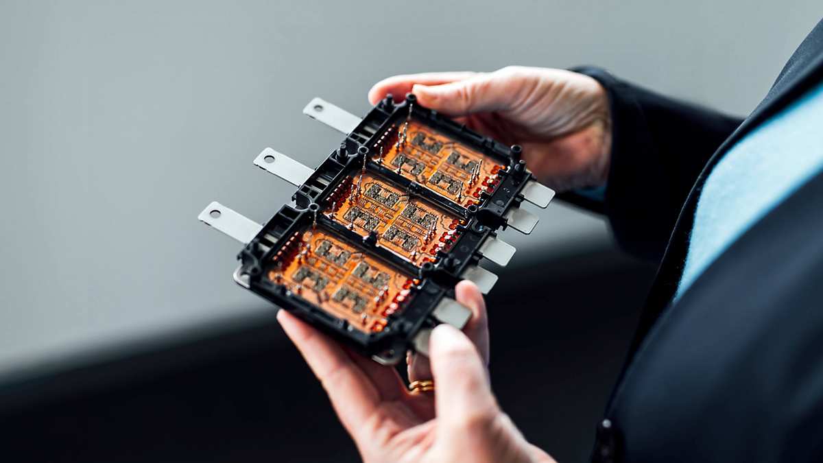 Close-up of an Infineon hybrid pack held in both hands of a woman wearing a blue blazer. Opened rectangular Hybrid Pack with a black casing and multiple metallic contact pins on the top and bottom side. Three large copper areas connected by numerous fine wires and contact pins. The module reveals the complex internal structure of power electronics used in hybrid and electric vehicles.